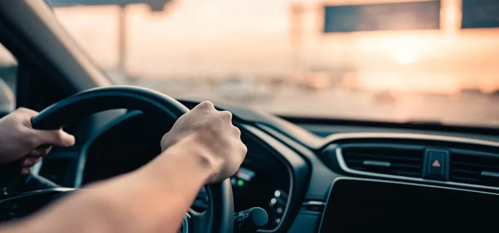 Detalhe das mãos no volante de um carro ao pôr do sol, ilustrando o uso e os benefícios da direção assistida.