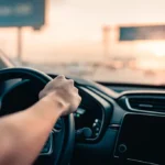 Detalhe das mãos no volante de um carro ao pôr do sol, ilustrando o uso e os benefícios da direção assistida.