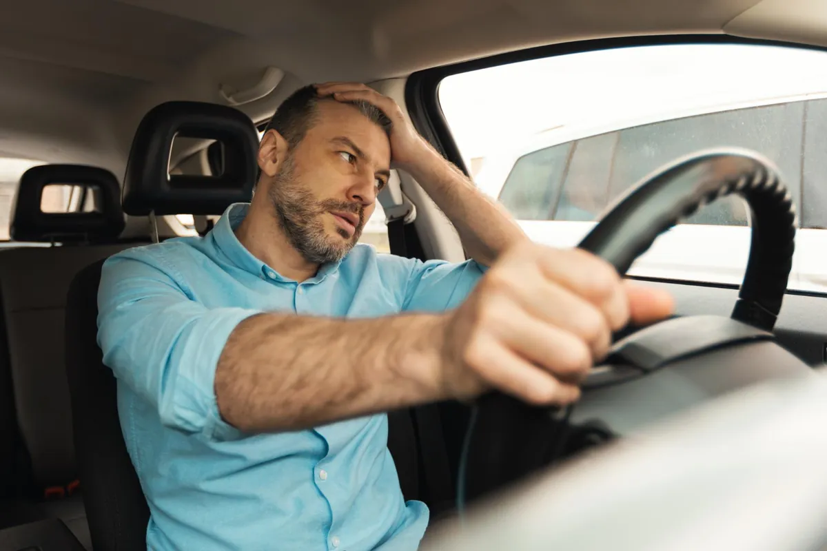 Sintomas de medo de dirigir Homem com a mão na cabeça, olhar tenso, segurando o volante do carro, demonstrando medo de dirigir