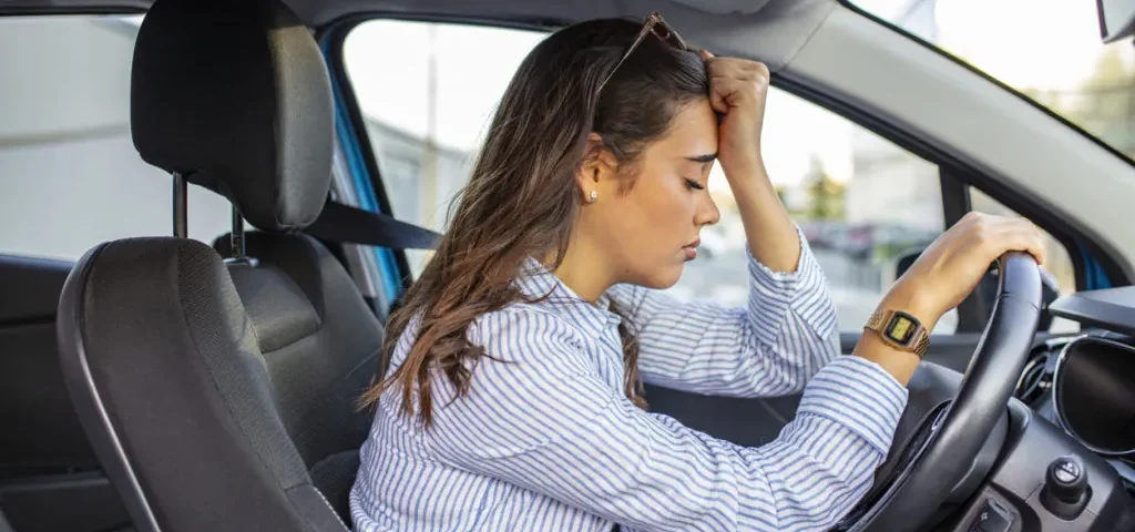 Mulher com a mão na testa, aparentando tensão, sentada no banco do motorista com a outra mão no volante, expressando medo de dirigir