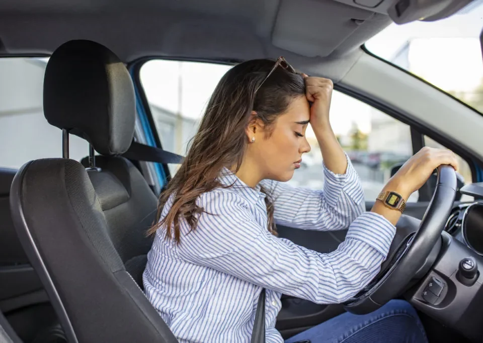 Mulher com a mão na testa, aparentando tensão, sentada no banco do motorista com a outra mão no volante, expressando medo de dirigir
