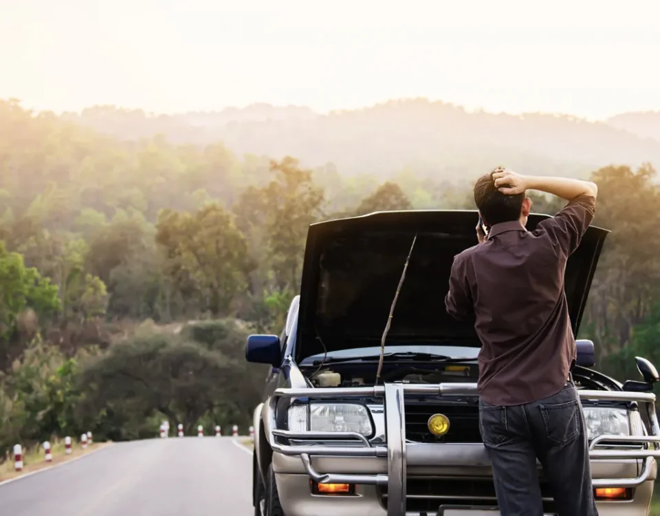 Homem de costas com a mão na cabeça e telefone no ouvido diante de um carro com capô aberto em estrada, representando uma pane elétrica no carro